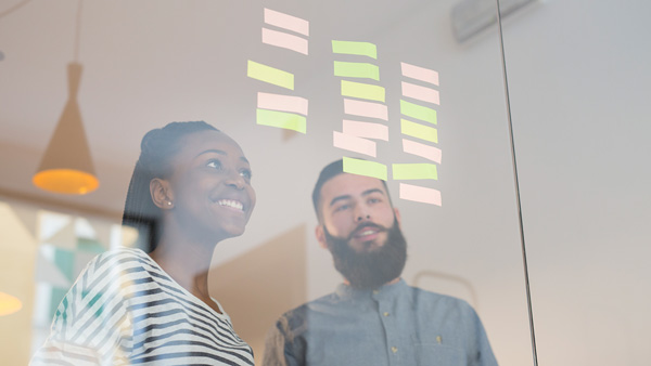 A pair of students smiling as they review ideas on post-it notes stuck to a glass partition