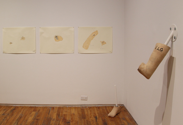 A corner of the Tangled Art + Disability gallery with drawings on the back wall, a foot sculpture on the floor, and an elbow joint sculpture mounted on the side wall.