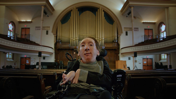 Composer Ari Kinarthy sitting in front of a large concert hall organ.