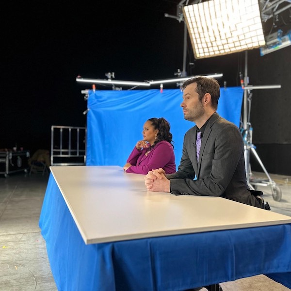 Gaitrie Persaud-Killings as news anchor Arianna Salara and Graham Kent as news anchor Grant Gewürtztraminer are shown sitting at a television news desk.