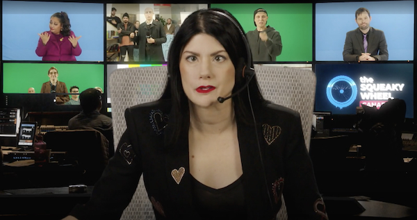 A news anchor with a headset microphone addresses the camera. There is an array of screens behind her.