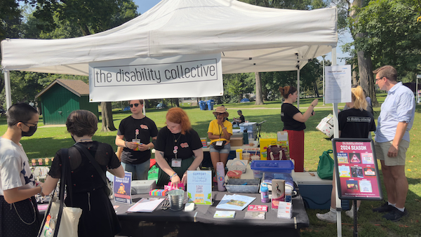 A photograph of The Disability Collective booth at the Disability Artisan Market. It is outdoors at a city park. There are a number of people behind the booth and a table with flyers and information about the organization and the market.