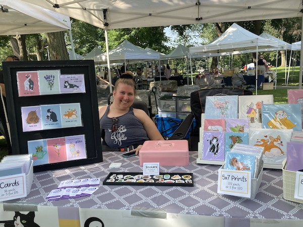A photograph of the artist Brooklyn Marx at her Disability Artisan Market booth. She is smiling behind a table displaying cards, buttons, and prints of her drawings.