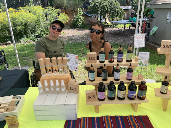 A photograph of the Bright Holler booth at the Disability Artisan Market. The two herbalists sit behind a table stocked with a selection of bottled tinctures and sprays.