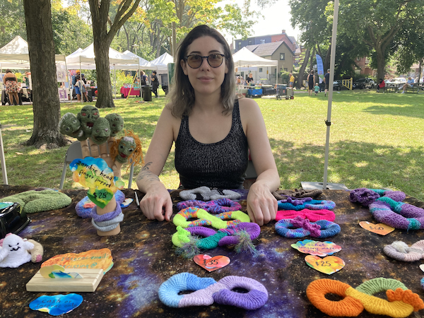 A photograph of the artist Kat Singer sitting behind their table at the Disability Artisan Market. On the table is a display of their crocheted bead mazes in a variety of bright colours.