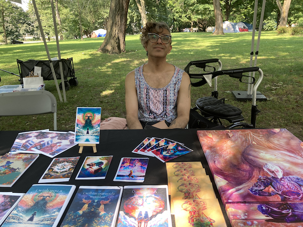 A photograph of the artist and writer Parvati the Art Pixie at the Disability Art Market. She sits behind a table displaying a selection of her artwork and zines.