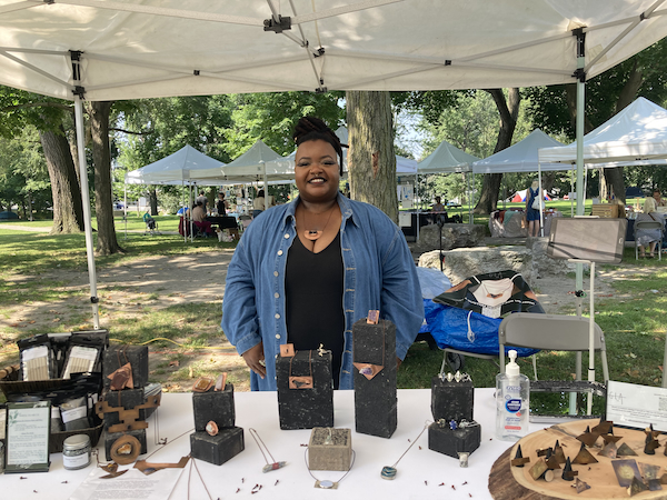 A photograph of Priscilla from Good Look Artistry standing behind her table at the Disability Artisan Market. The table displays a selection of her metal and stone jewellery.