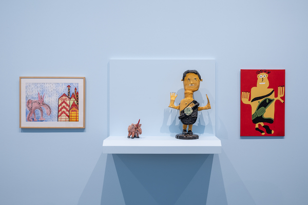 A photograph of two of Aaron Harvey's drawings and two of Boyd Dymchuk's sculptures in the Art Gallery of Alberta. The drawings are mounted on the wall and the sculptures are displayed on a shelf and in a glass cabinet.