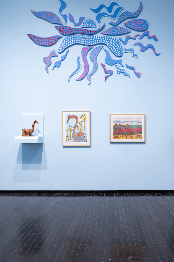 A photograph of a wood wall sculpture depicting blue flames, two drawings, and a small sculpture displayed on a shelf in the Art Gallery of Alberta.