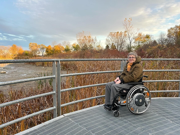 A photograph of Kim Fullerton, seated in her wheelchair and smiling at the camera, on a boardwalk overlooking a marsh on a autumn day.