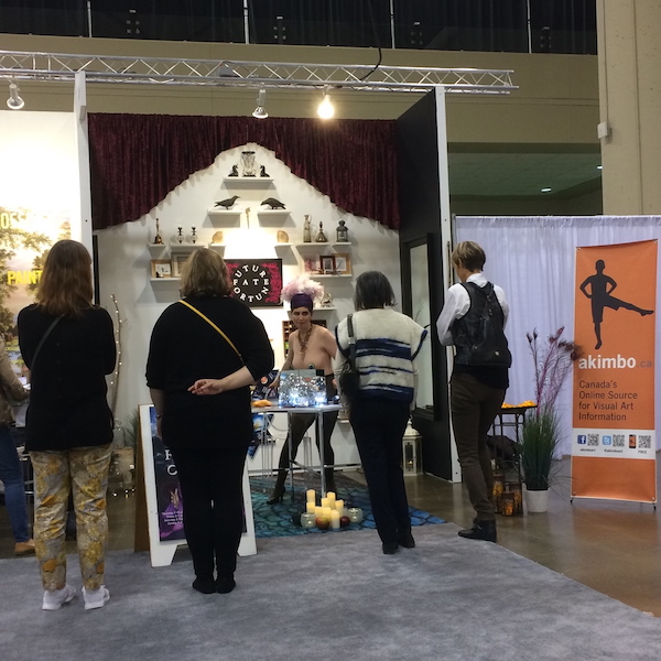 A photograph of the Akimbo booth at the Art Toronto art fair. The booth features the performance artist Jess Dobkin dressed up like an oracle. There are four visitors watching her.