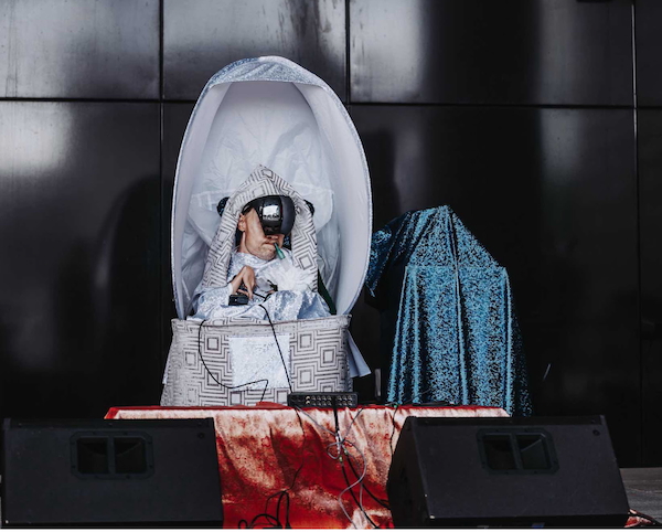 Bucko (Chris Binkowski) on stage, draped in patterned cloths and wearing a mirrored mask performs his music.