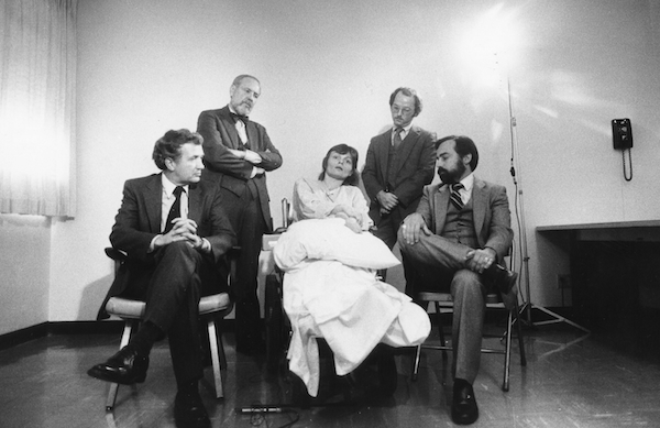 A black-and-white archival image of Elizabeth Bouvia, center, in her wheelchair. She holds a pillow in her lap, and gazes ahead — mouth agape and head slightly to the side. Around her, four male lawyers in suits are staged around her, staring at her seriously. Behind her to the left, one of the lawyers crosses his arms at his chest while looking down at her, as if to scold her. 