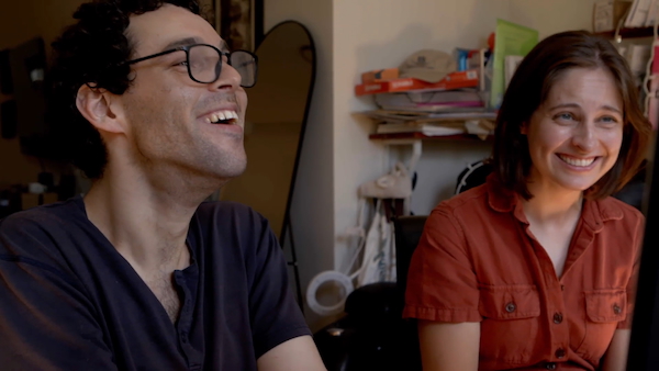 Director Reid Davenport, a white man with curly, short brown hair and glasses, and Producer Colleen Cassingham, a white woman with a brown bob wearing an orange jumpsuit, laugh together as they look toward a computer screen, which is just out of the shot. In the background, is an apartment with a full length mirror and an array of books on a shelf. 