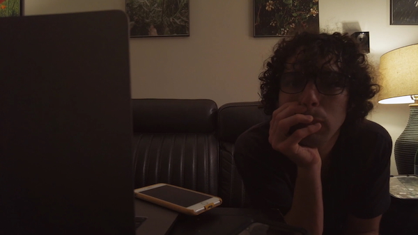 Director Reid Davenport thinking on his couch, with his head resting in his hand. He is a white man with curly brown hair and glasses, and he looks at the camera. To his left in the foreground is a laptop and a phone on a side table. 