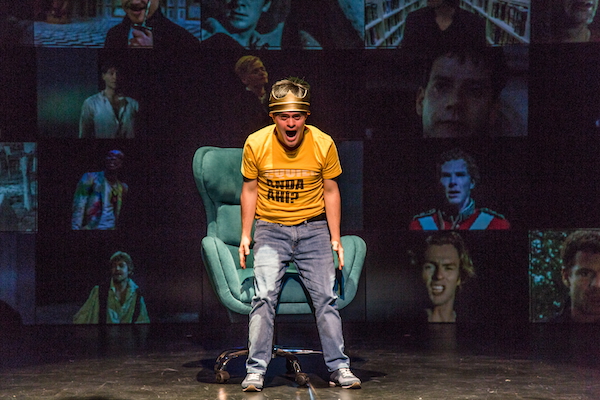 Actor Jaime Cruz is dressed in jeans, a yellow t-shirt and a crown. He is screaming and standing in front of a wall of monitors featuring various actors portraying Hamlet.