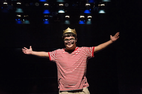 Actor Alvaro Toledo, dressed in a red and white striped t-shirt and wearing a crown, stands smiling with his hands held out wide.