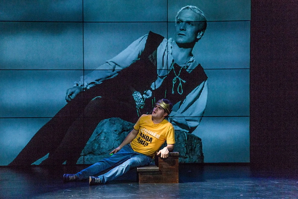 Actor Jaime Cruz is dressed in jeans, a yellow t-shirt and a crown. He is reclining on a step in front of a wall of monitors displaying Laurence Olivier playing Hamlet.