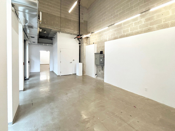 Interior of Gallery A: daylight from left (windows out of frame); right wall with plinth, closed bathroom door, and open door to Gallery B.