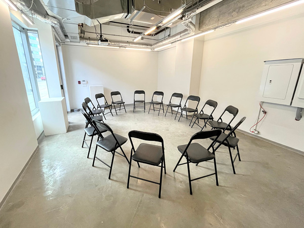 Interior of Gallery B: 15 black chairs form a circle in center; bright daylight from back-left window near two white plinths.