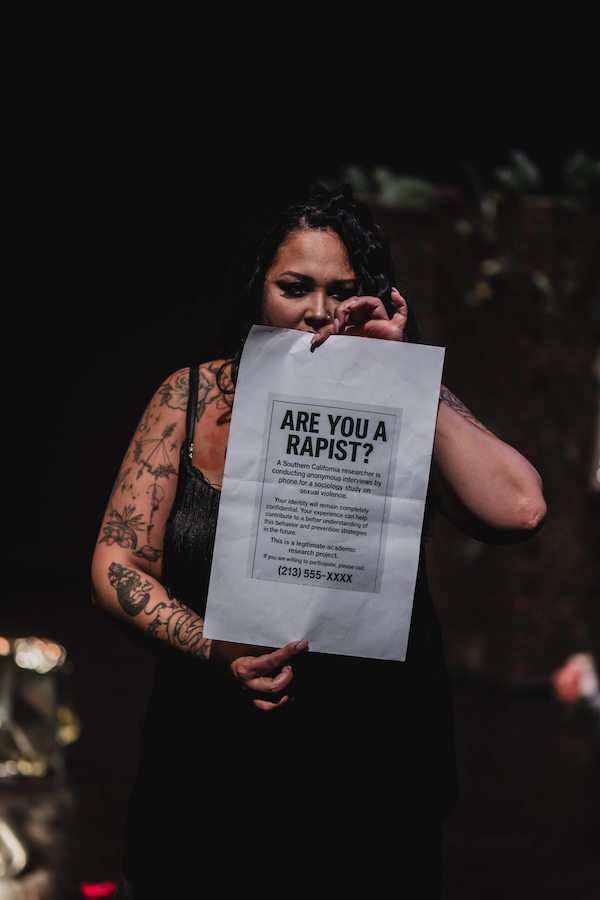 A woman on stage holds a copy of a poster in front of her. The poster reads, "Are you a rapist?"