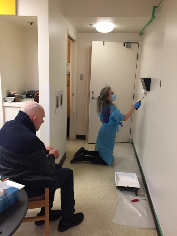 A woman is kneeling and painting a wall with a roller while a man sits and watches her.