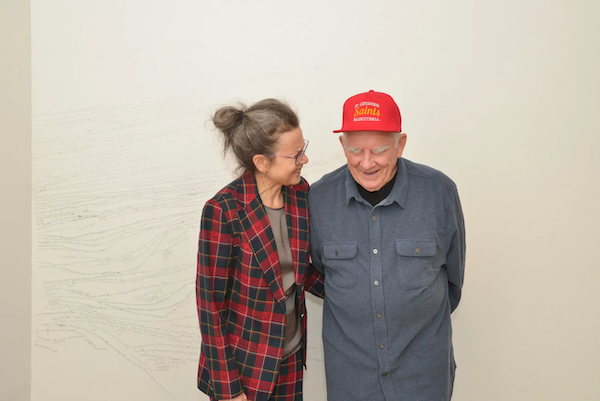 A woman in a plaid jacket and a man in a grey shirt and red hat smile at each other in front of a white wall with small writing on it.