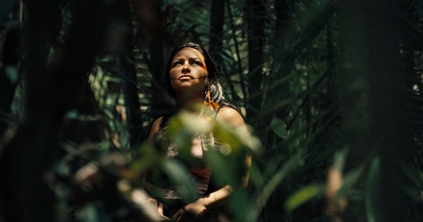 Production still from “Yanuni” showing a woman standing in a forest, lit by dappled sunlight