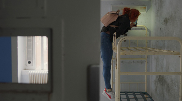 Production still from “Holloway” showing a woman on a bunk bed using her phone flashlight to illuminate something on the wall