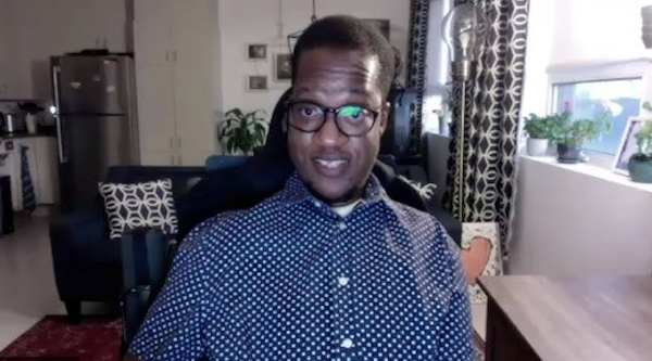 A Black man is seated in his wheelchair in his living room. He is looking directly at the camera.