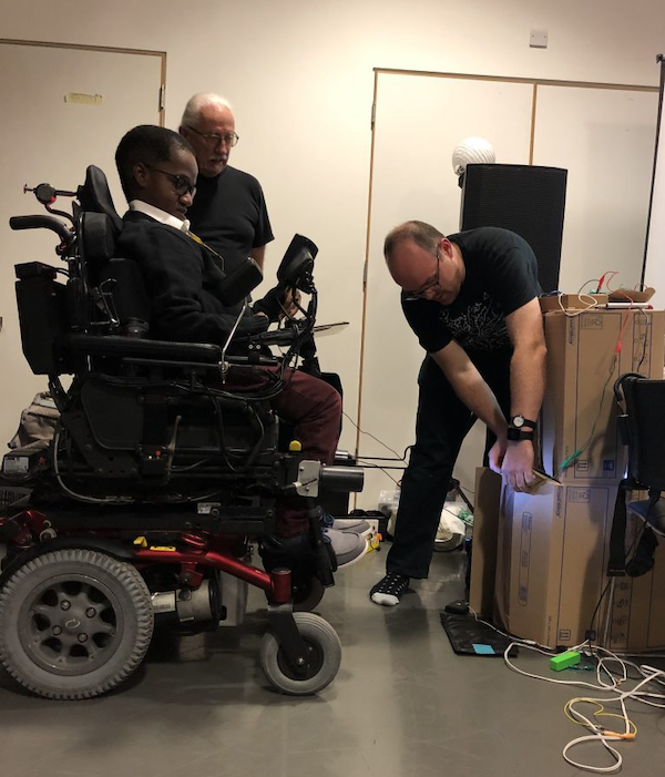 Gift Tshuma is sitting in his wheelchair and facing another man who is working on some electronic gear attached to a cardboard box.