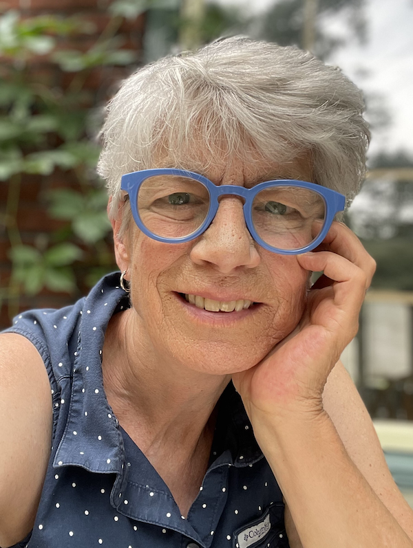 This portrait of Nancy Huggett depicts her smiling as she rests her head on one hand. She is wearing blue-framed glasses.