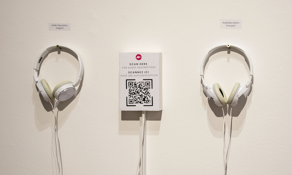 Two white headphones hang on a gallery wall beside a QR code sign labeled for English and French audio description. Small labels above each headset indicate language options for accessing exhibition audio description.
