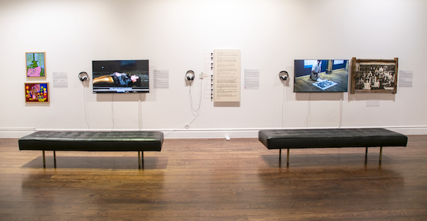A wide shot of the gallery wall in Crip Canada: A Decade of Political Engagement. From left to right, two small colourful paintings by Alexander Stuber at left, documentation of Vanessa Dion Fletcher’s Finding Language performance on a TV monitor, a Braille canvas with copper-line touchboard interface by Olivia Brouwer at centre, and Michel Dumont’s repaired ceramic tile mosaic classroom photograph at right. Monitors with headphones beside the works provide audio components. Two black benches sit in the foreground for seated viewing.
