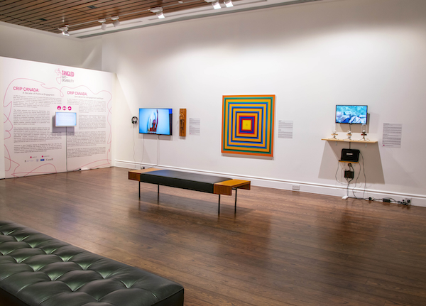 An installation view of Crip Canada: A Decade of Political Engagement shows the exhibition introduction wall text at left, a monitor with headphones and Persimmon Blackbridge’s soft touch, Peter Owusu-Ansah’s square concentric-colour piece at centre, and David Bobier’s vibrotactile sound installation with small glass domes at right. A long bench sits in the foreground facing the works.