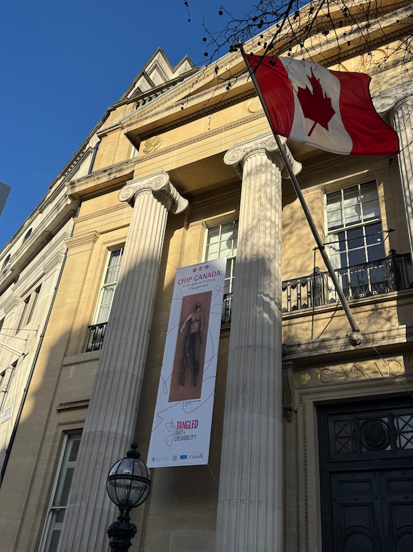 A vertical banner for Crip Canada: A Decade of Political Engagement hangs between tall stone columns at the entrance to Canada House in London. The building is a neoclassical building with a Canadian flag extending outward from the façade above the entrance.