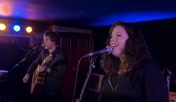 A guitarist and a singer performing in a small venue. They are both singing into microphones and lit up by stage lights.