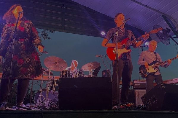 A colour photograph of the four members of the musical group The LH Express playing a concert. A singer and a guitarist are in the foreground. A bassist and a drummer are in the background.