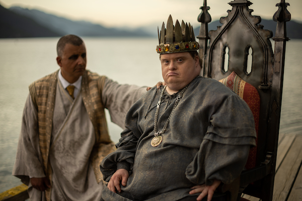 Niall McNeill, playing King Arthur, sits on his throne with Merlin, played by Marcus Youssef, crouched behind him. A lake is in the background.