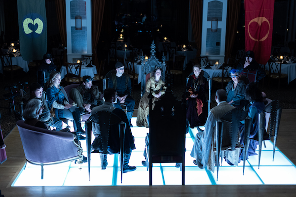 The cast of King Arthur's Night, dressed in formal costumes, sit in a circle in a darkly lit room.
