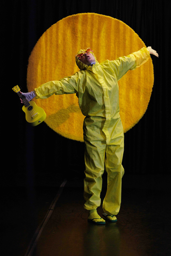 Sara Porter is dressed in yellow coveralls with a hood on and sunglasses. She holds her arms outstretched, with a ukulele in one hand, and sunglasses on. She is standing in front of a cut-out of the sun.