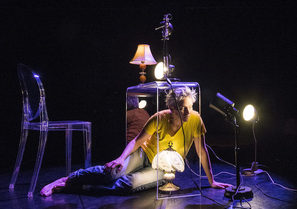 Sarah Porter is dressed in jeans and a yellow t-shirt. She is sitting on the floor under a plexiglass desk, illuminated by a couple lamps.
