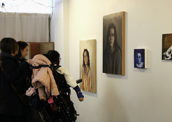 A photograph of a couple people looking at three paintings of human figures on a gallery wall.