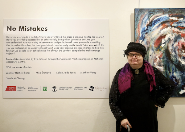 A photograph of the exhibition curator standing in front of the exhibition wall text and a painting.