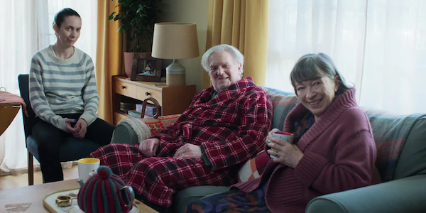 An elderly man and woman sit, smiling, on a couch. There is a younger woman on a chair beside them. This is a scene from the movie Tender Loving Care.