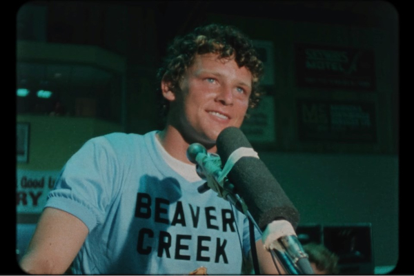 In a still from the documentary Run Terry Run, Terry Fox is smiling and standing in front of a microphone at a press conference.